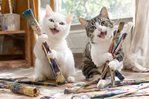 白猫とキジ白猫が絵の具のついたたくさんの筆を持っている。周りにも筆が転がっている。楽しそう！